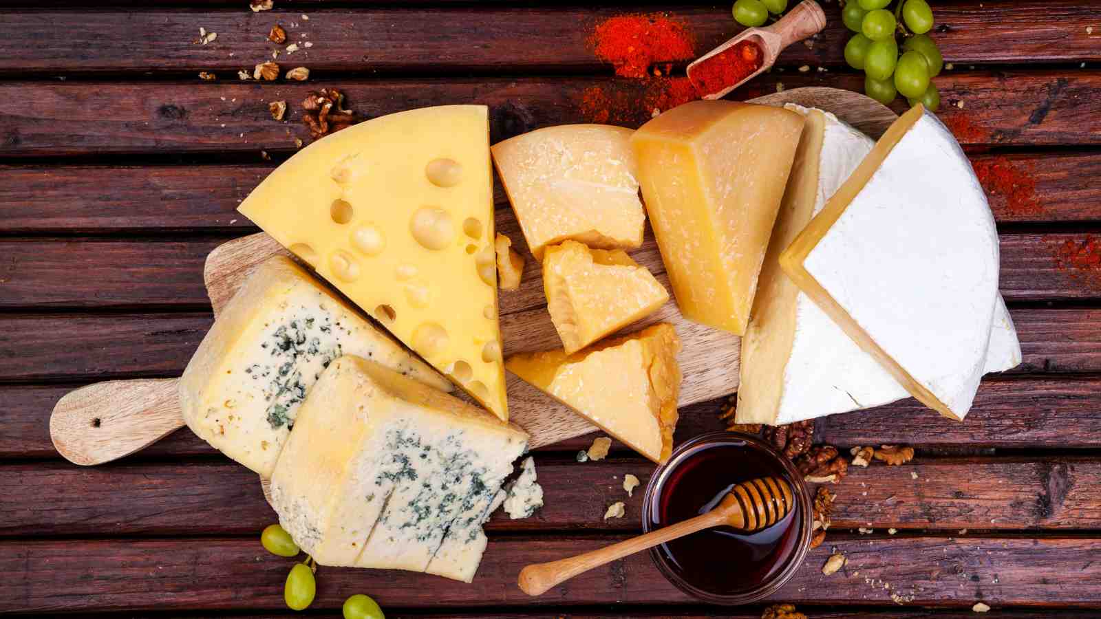 Assorted cheeses including blue, Swiss, and Brie are displayed on a wooden board with grapes, nuts, honey, and a dusting of paprika.