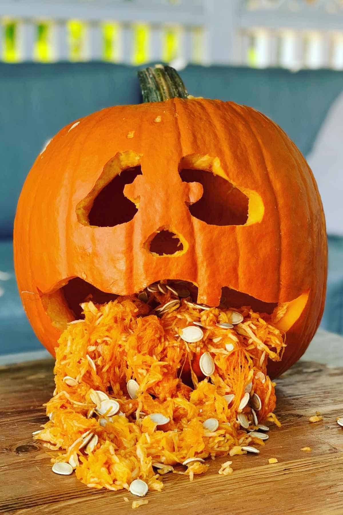 Carved pumpkin with a face appears to be vomiting its insides, with seeds and pulp spilling onto a wooden surface.