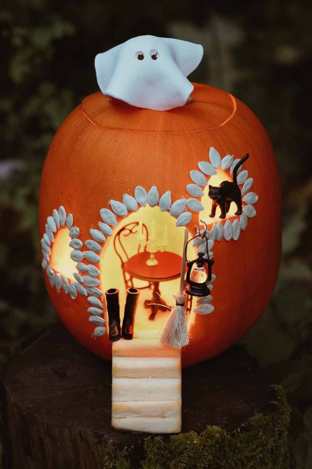 A carved pumpkin with a lit interior displays a miniature room scene, decorated with a ghost on top and a black cat.