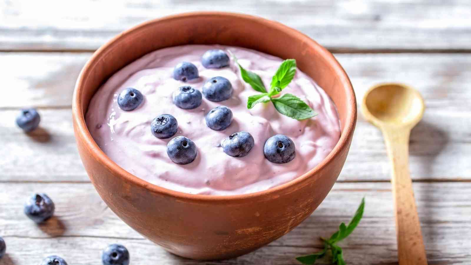 A wooden bowl of blueberry yogurt garnished with fresh blueberries and a sprig of mint, placed on a wooden surface beside a wooden spoon.