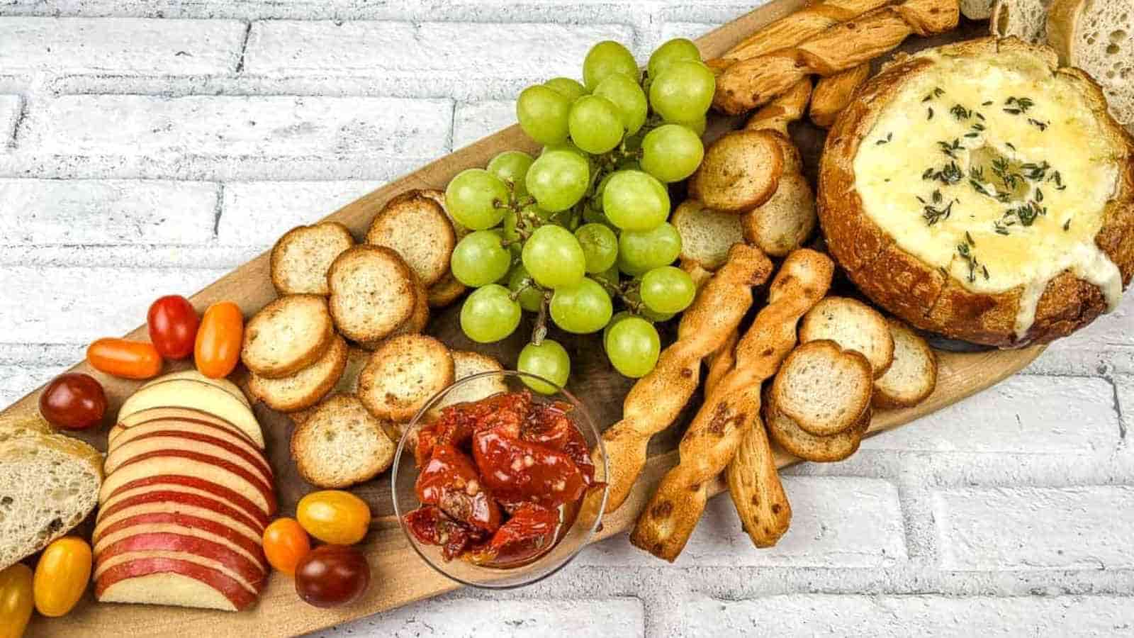 A wooden platter with baked brie in a bowl, cheese, crackers, grapes and bread.