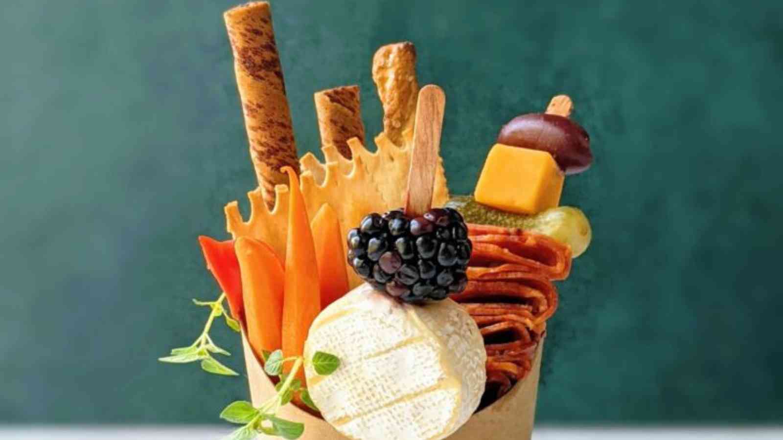 A paper charcuterie cup holding a variety of cheeses, pretzels and meats.