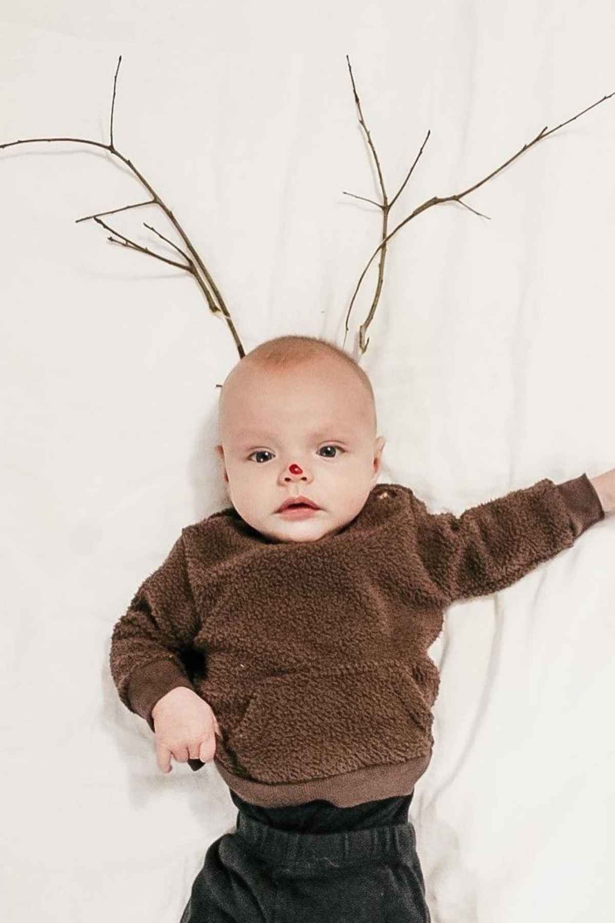 Baby dressed in a brown sweater with red nose makeup, lying on a white blanket with branches arranged to resemble antlers.