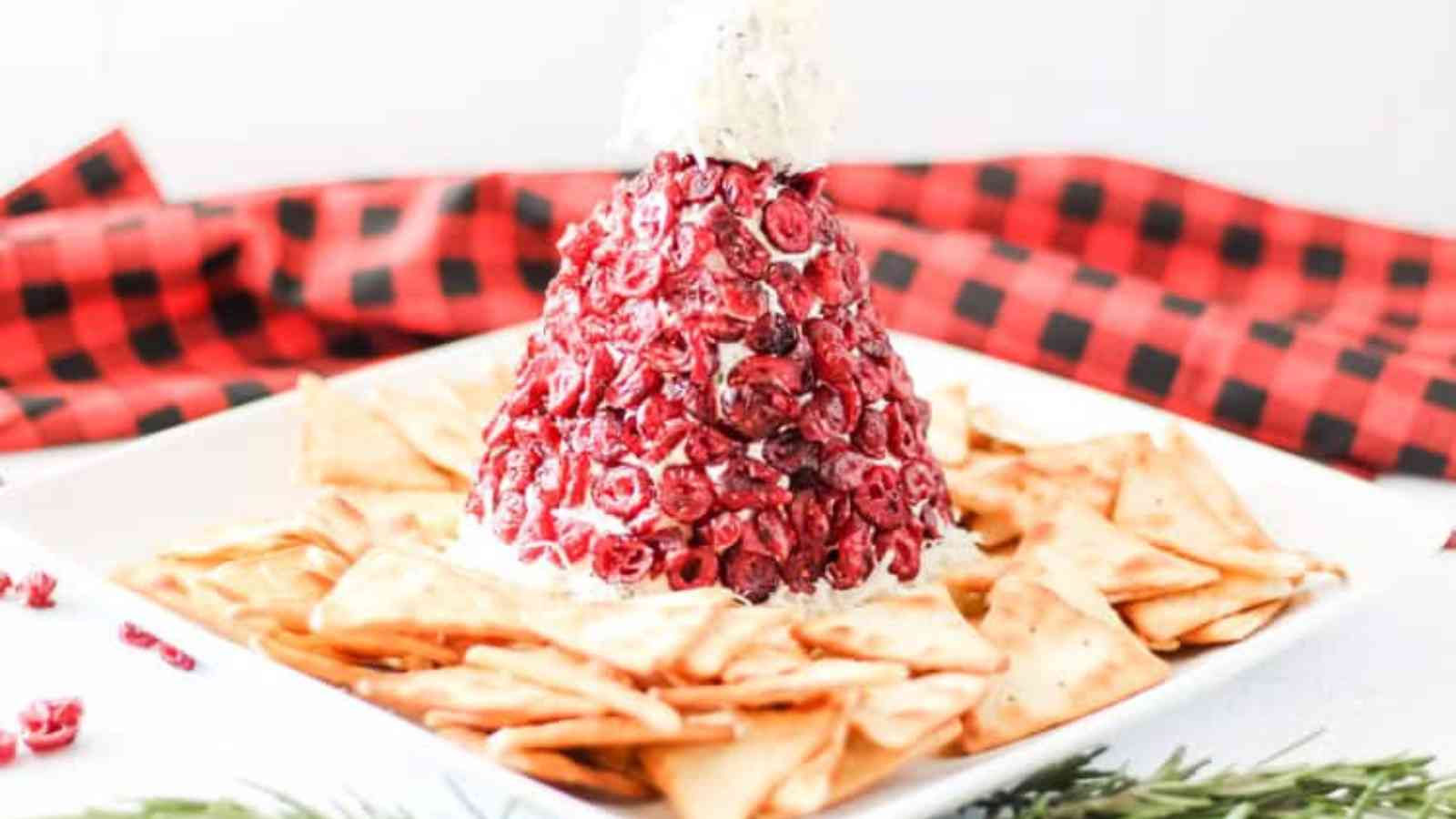 A colorful layered cheese ball shaped like Santa's hat, topped with red cranberries, surrounded by crackers on a white plate.
