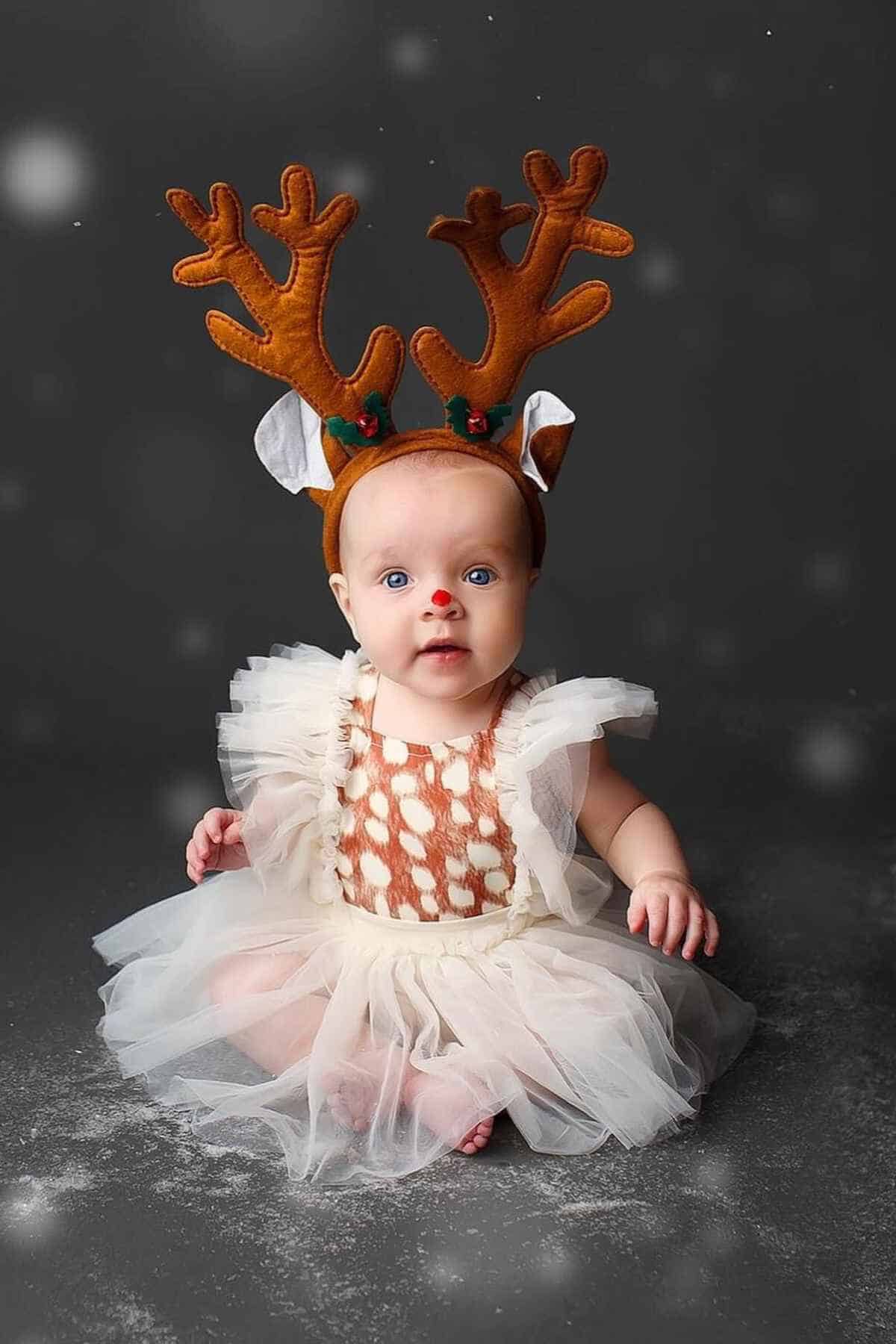 A baby wearing a reindeer antler headband and a white tulle dress sits on a snowy surface against a dark background.