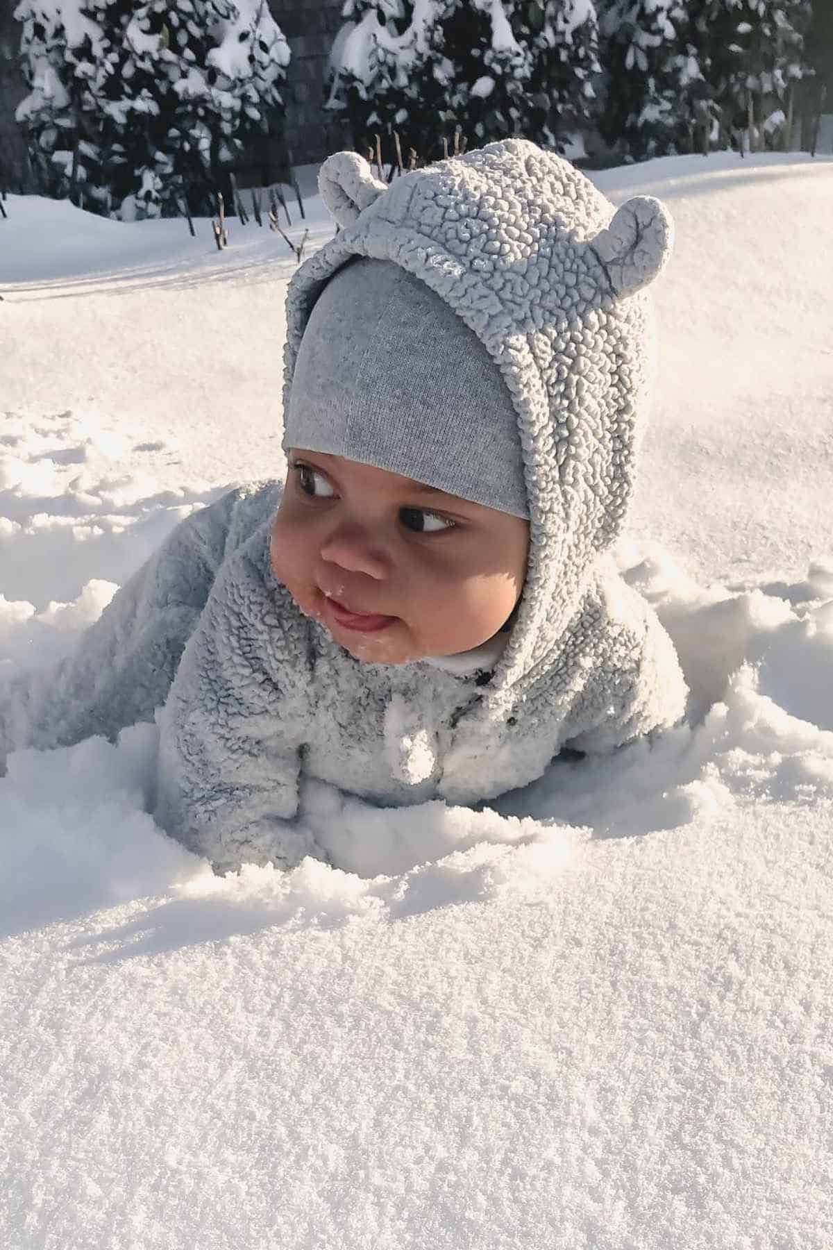 Baby in a fluffy gray outfit and hat lies on the snow, surrounded by a snowy landscape.