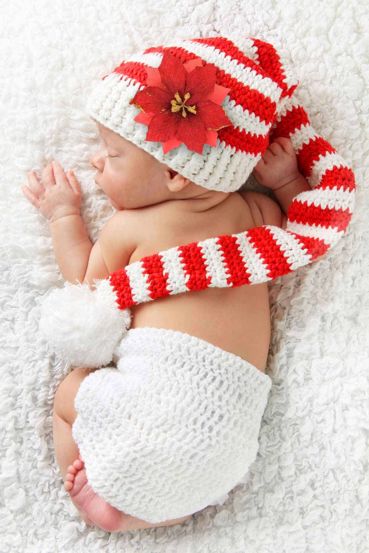 Newborn baby wearing a red and white striped hat with a flower, and a white crocheted diaper cover, lying on a fluffy white blanket.