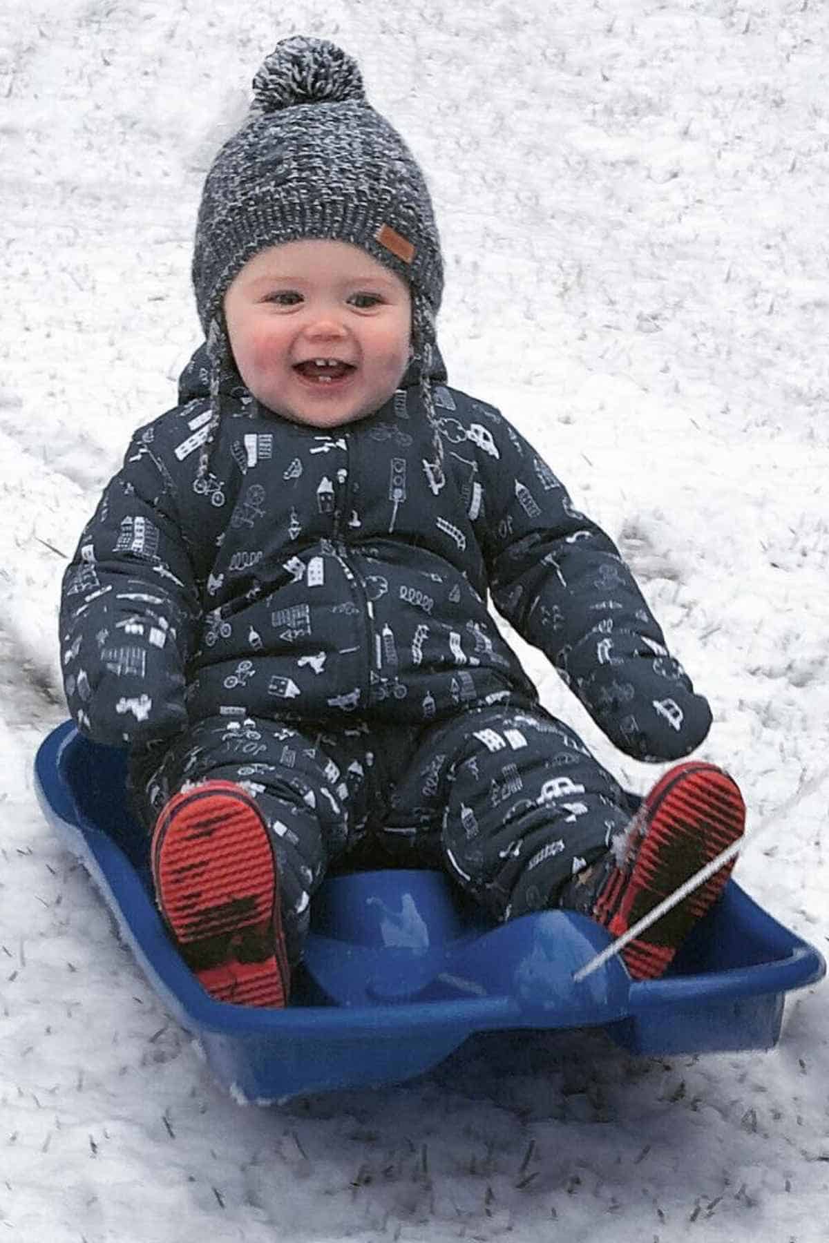 A smiling child in winter clothing sits on a blue sled in the snow.
