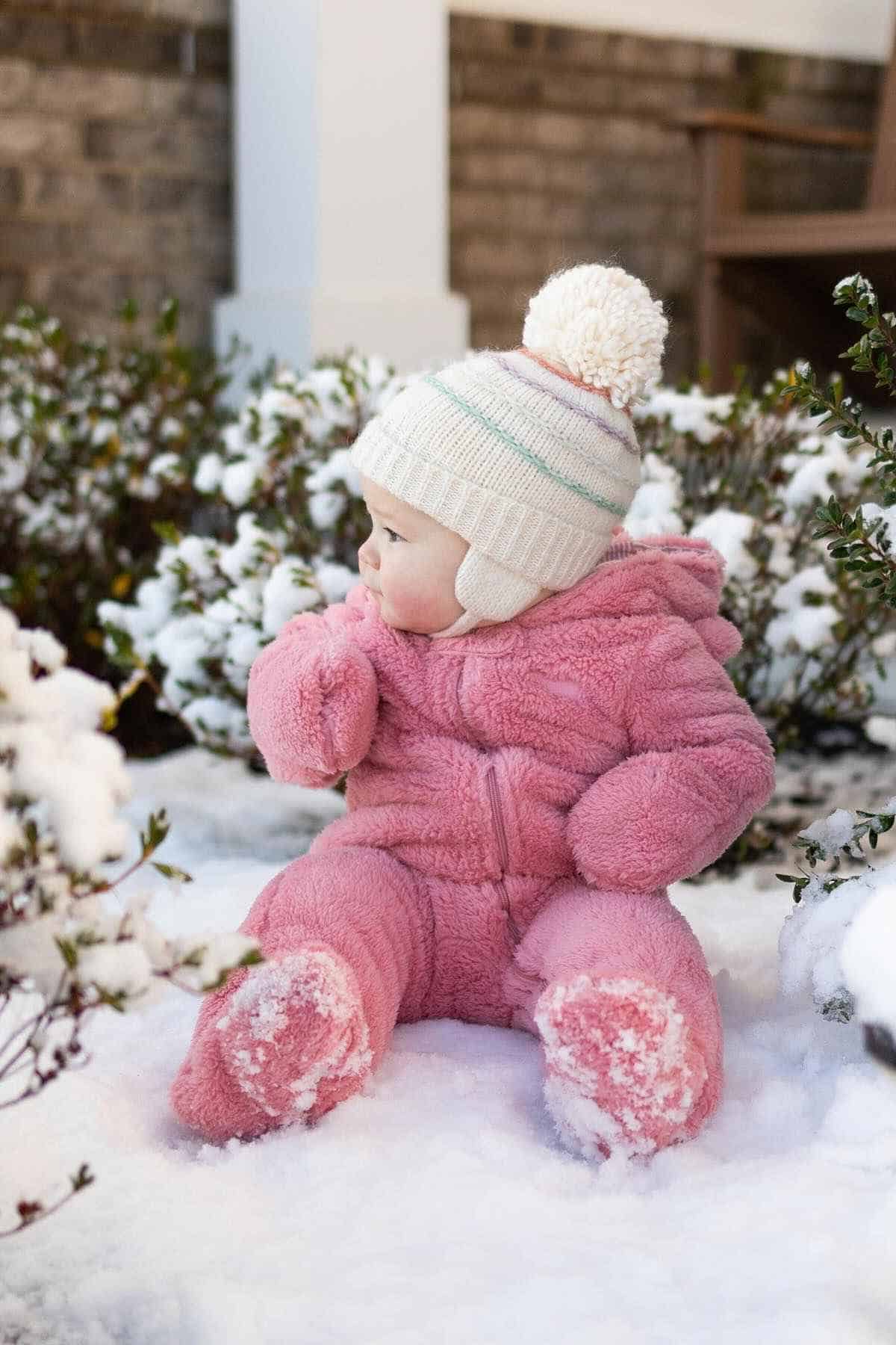 A baby in a pink snowsuit and white hat sits in the snow, surrounded by bushes.