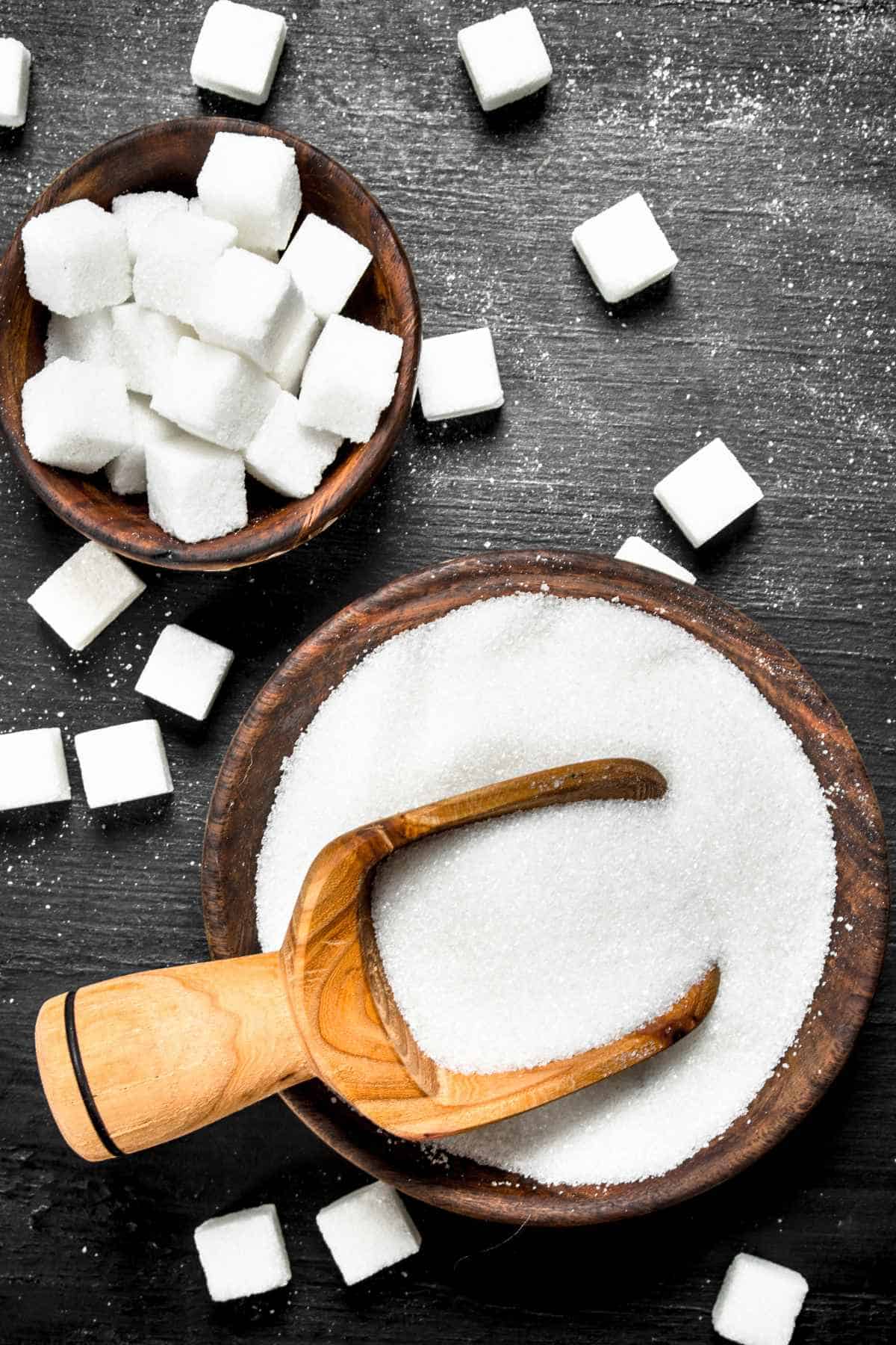 Wooden bowls filled with granulated sugar and sugar cubes rest on a dark surface.