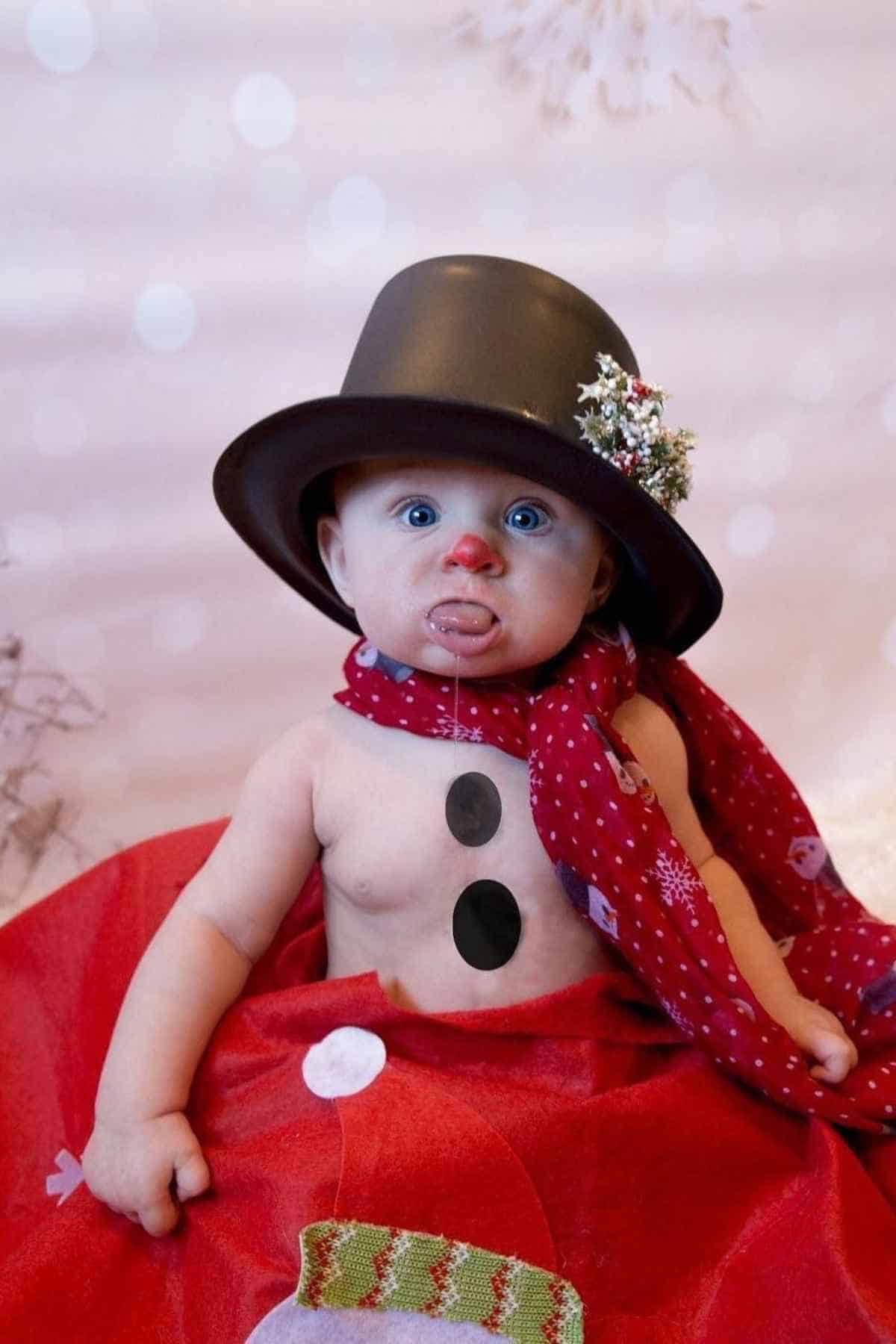 A baby dressed as a snowman, wearing a black hat with a flower, a red scarf, and has red on their nose.