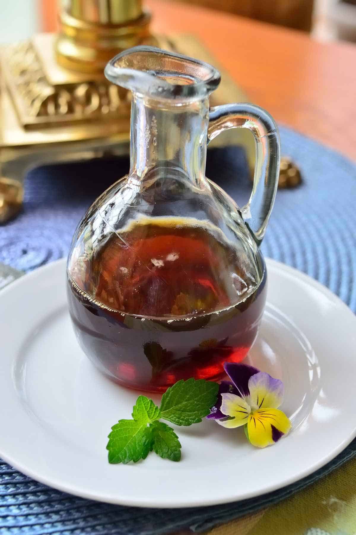 A glass pitcher with dark liquid sits on a white plate, accompanied by mint leaves and a yellow pansy flower.