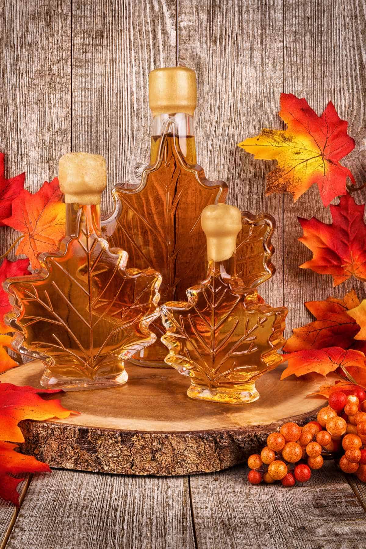Maple leaf-shaped bottles of syrup sit on a wooden surface, surrounded by autumn leaves and berries.