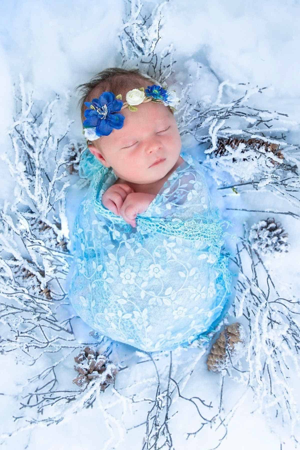 A sleeping baby wrapped in blue lace lies on a snowy surface, surrounded by frosty branches.