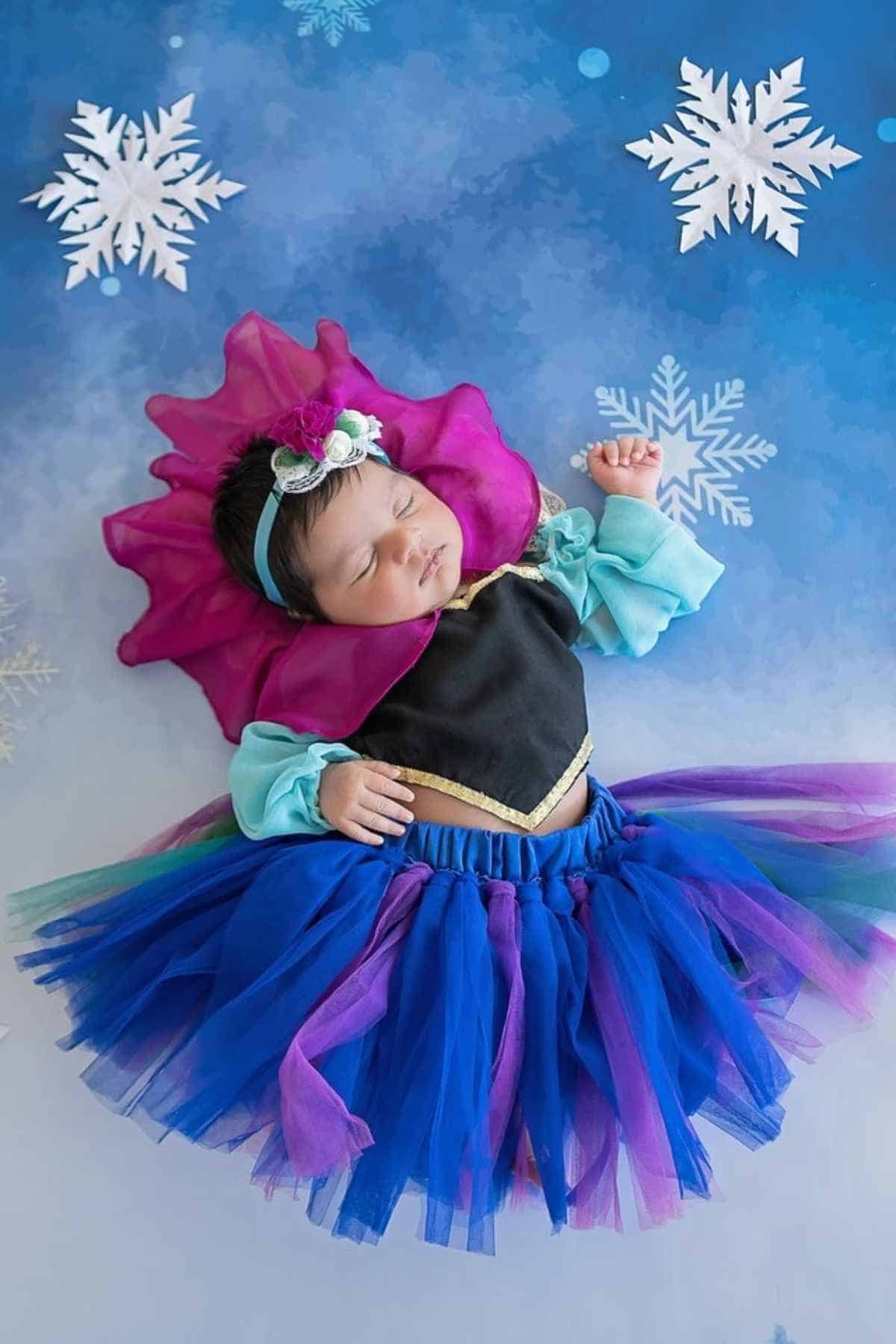 Newborn in a colorful costume lying on a blue background with snowflake decorations.