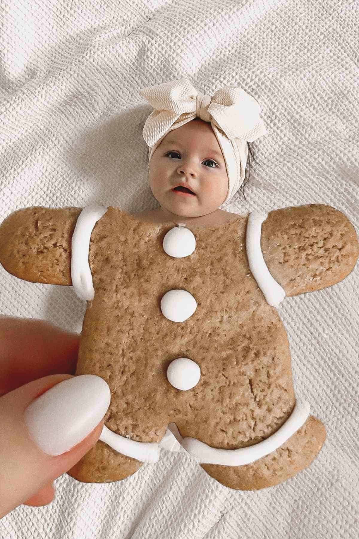 A baby with a bow headband is positioned behind a gingerbread cookie, creating the illusion of the baby having a gingerbread body.