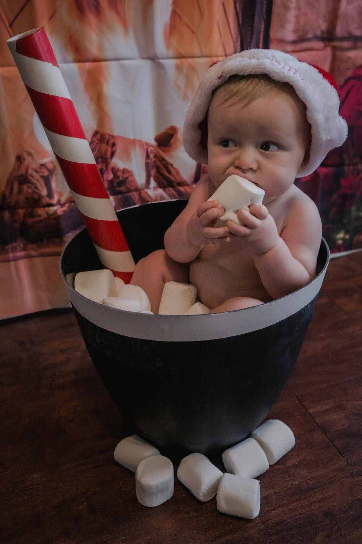 A baby wearing a Santa hat sits in a black bucket filled with marshmallows, holding a candy cane.