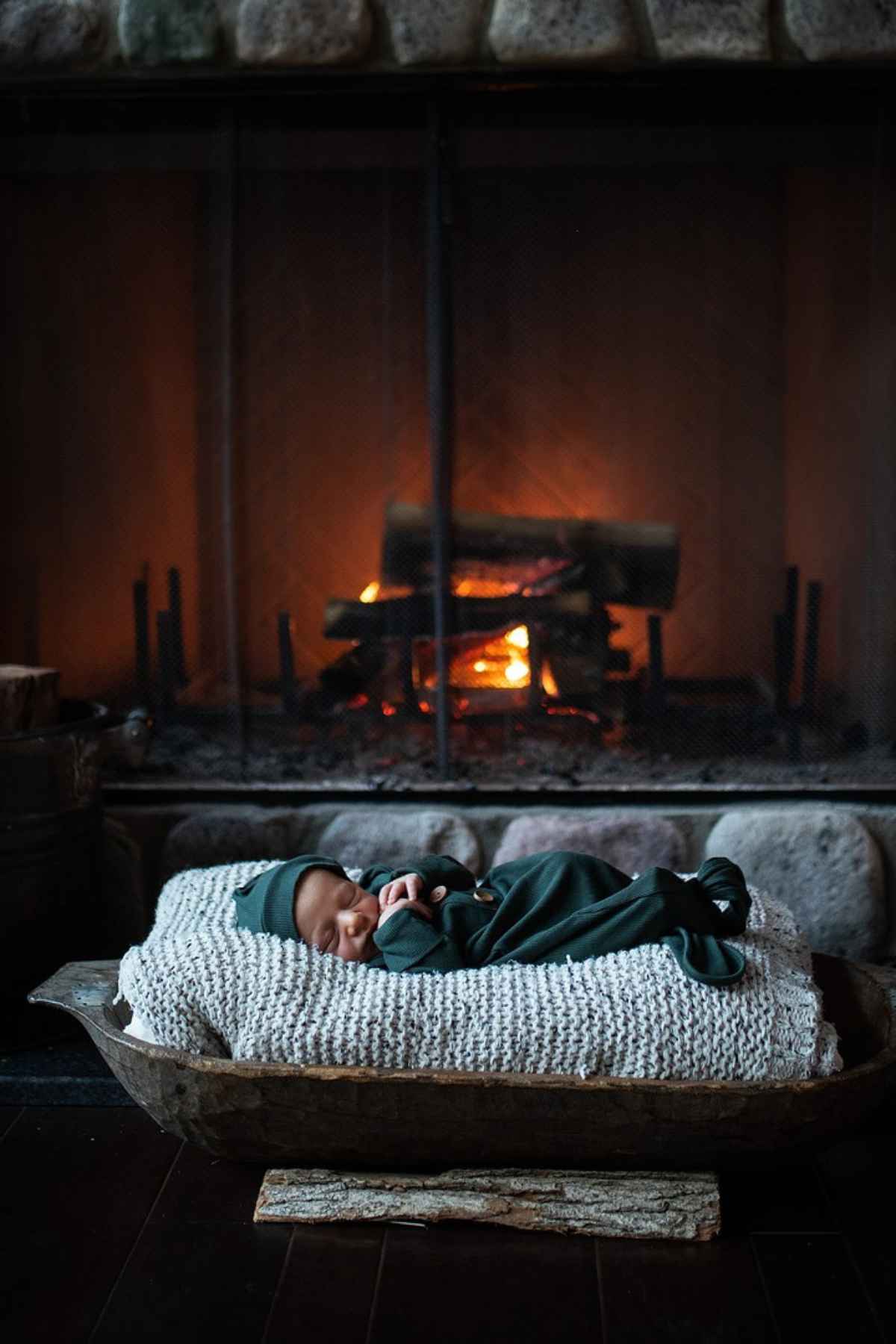 A baby in a dark outfit sleeps on a knit blanket, placed in a wooden trough in front of a lit fireplace.