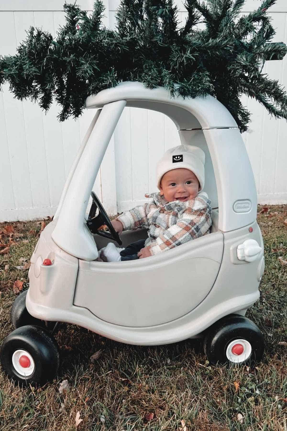 A baby in a toy car with a pretend Christmas tree on top, outdoors on grass.