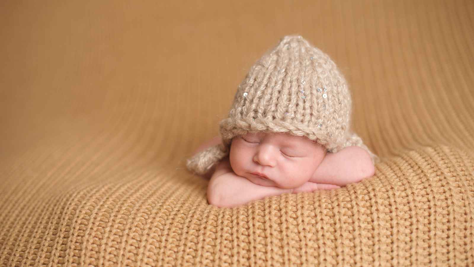 A sleeping baby rests peacefully on a knitted blanket, wearing a matching knitted hat.