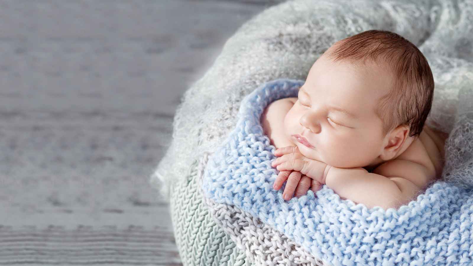 A newborn baby rests peacefully on a soft, blue knitted blanket, placed on a cushioned surface.