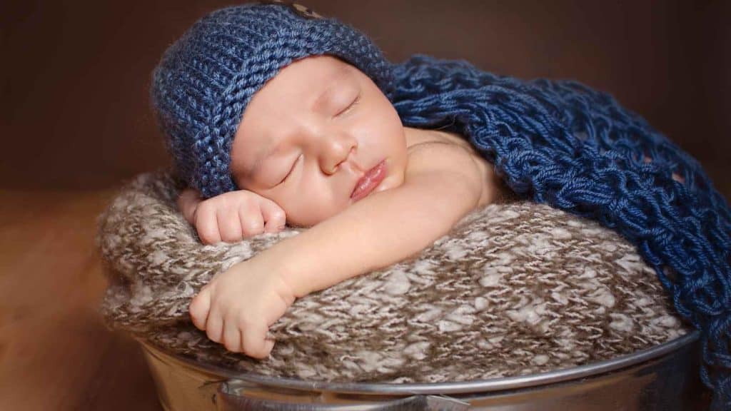 A sleeping baby, perhaps destined for one of those noble boy names, wears a blue knitted hat and blanket as he rests peacefully on a textured pillow inside a metal bucket.