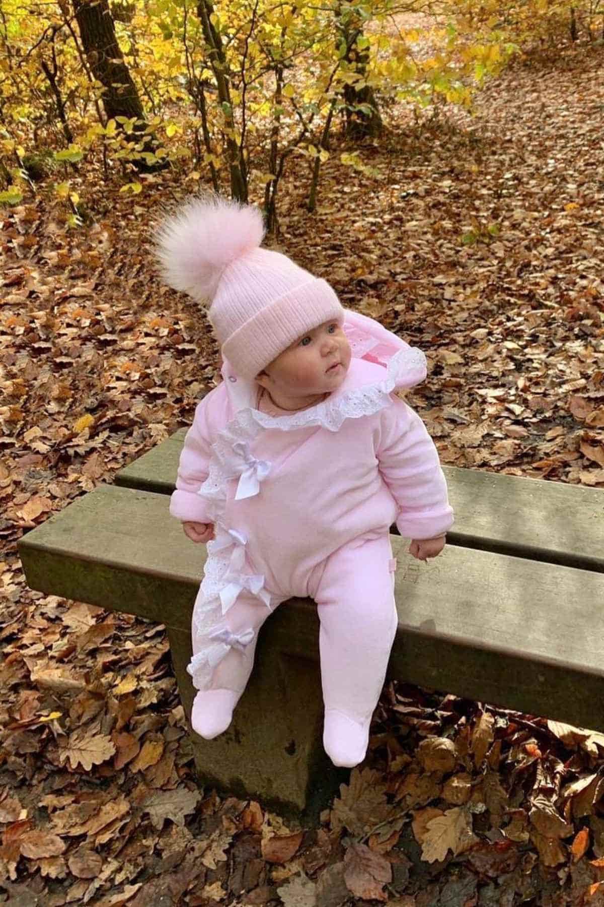 A baby in a pink outfit and fluffy hat sits on a wooden bench surrounded by autumn leaves.