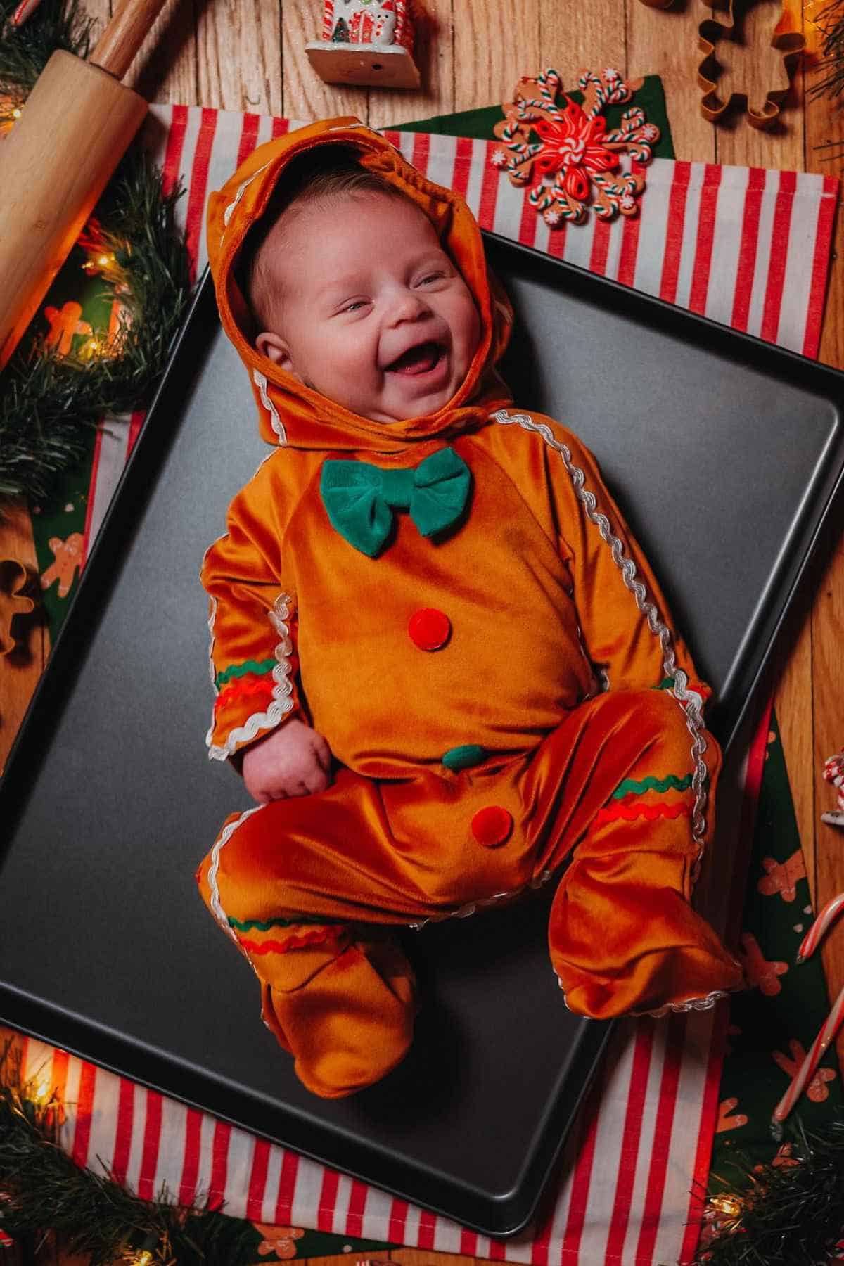 Baby in an orange gingerbread costume with a green bow, lying on a baking tray surrounded by festive decorations.