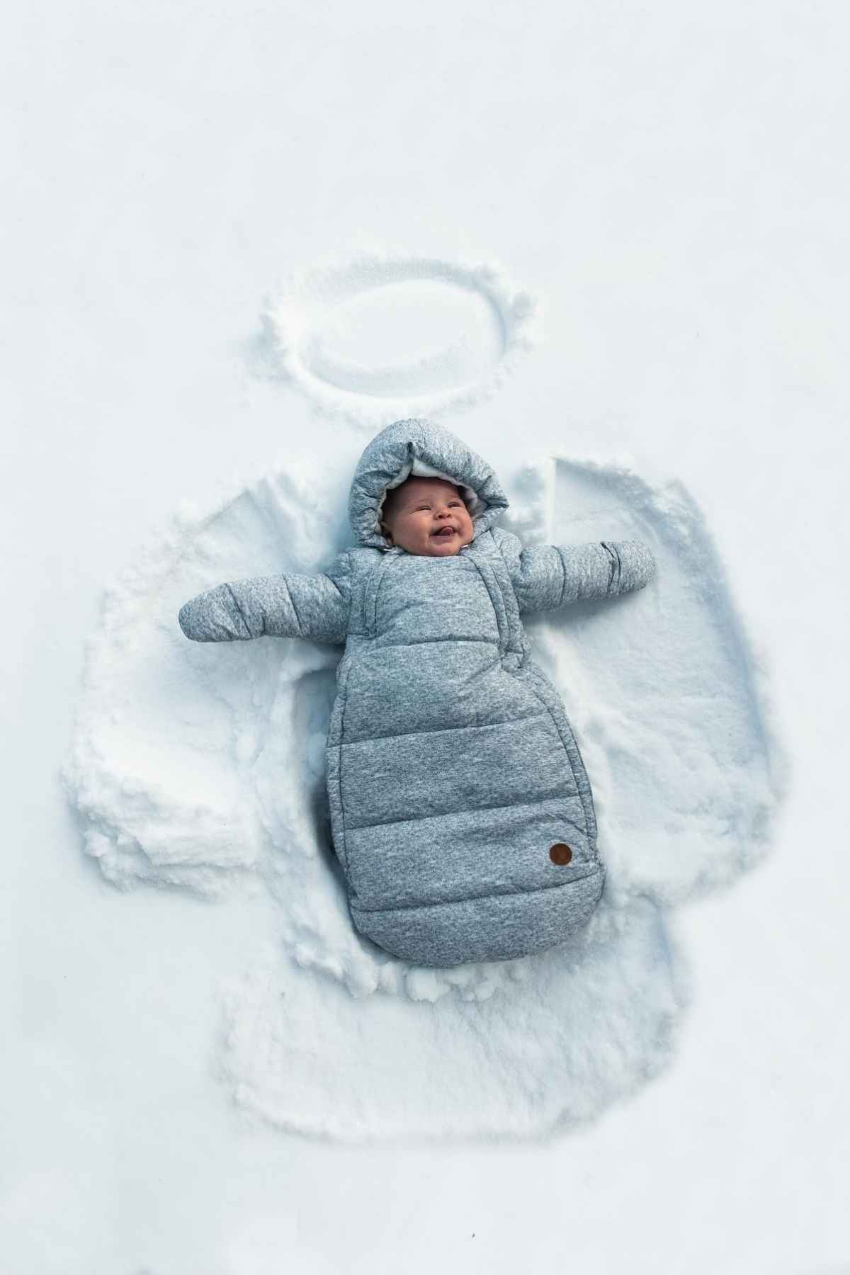 A baby in a gray snowsuit lies on the snow, making a snow angel.