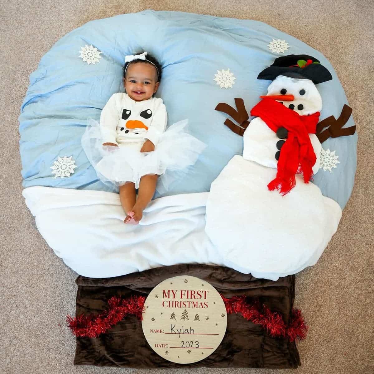 A baby in a snowman-themed setting, wearing a snowman outfit, lays on a blanket with a plush snowman.