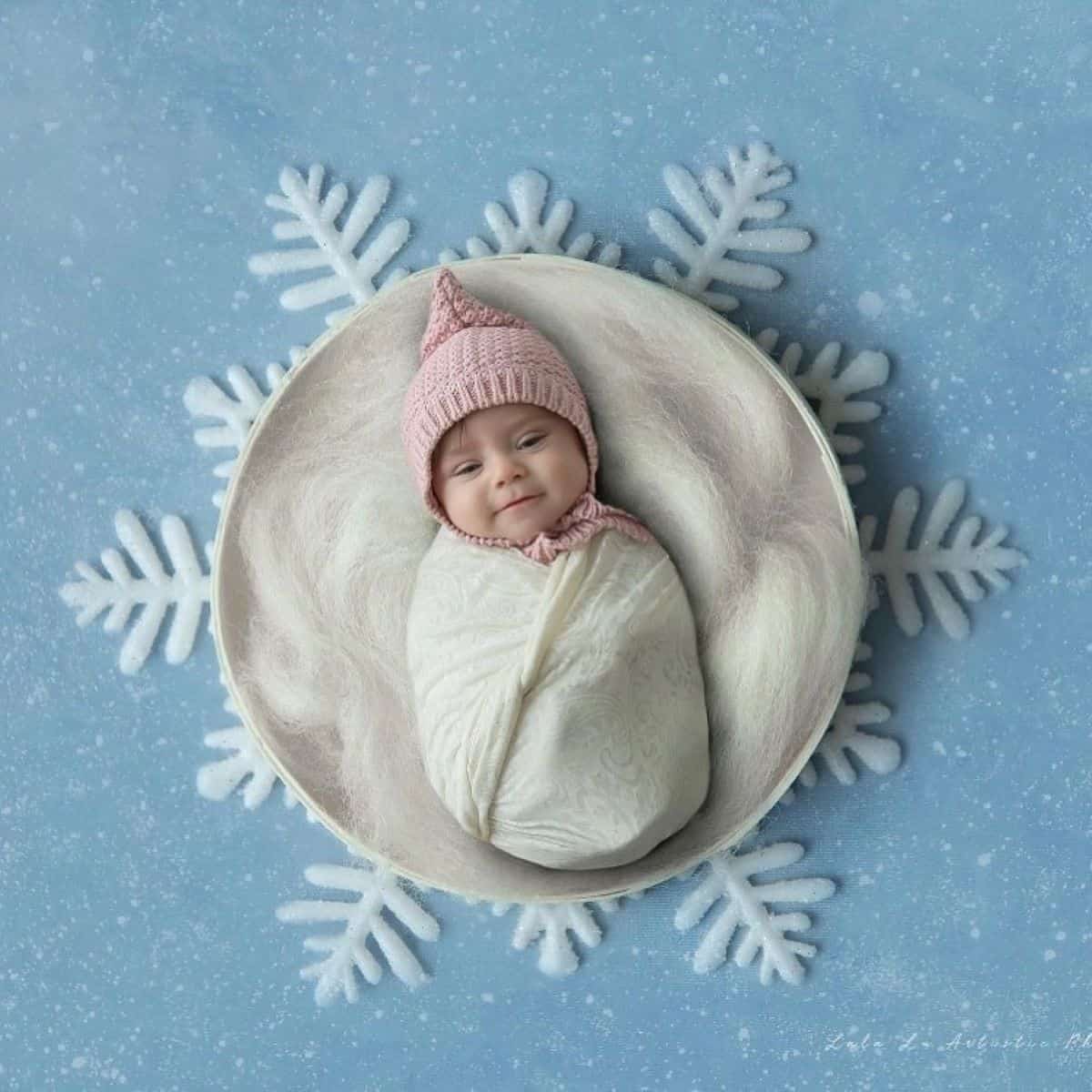 A baby in a pink hat is swaddled in a white blanket, lying on a circular cushion set against a blue background with large snowflake designs.