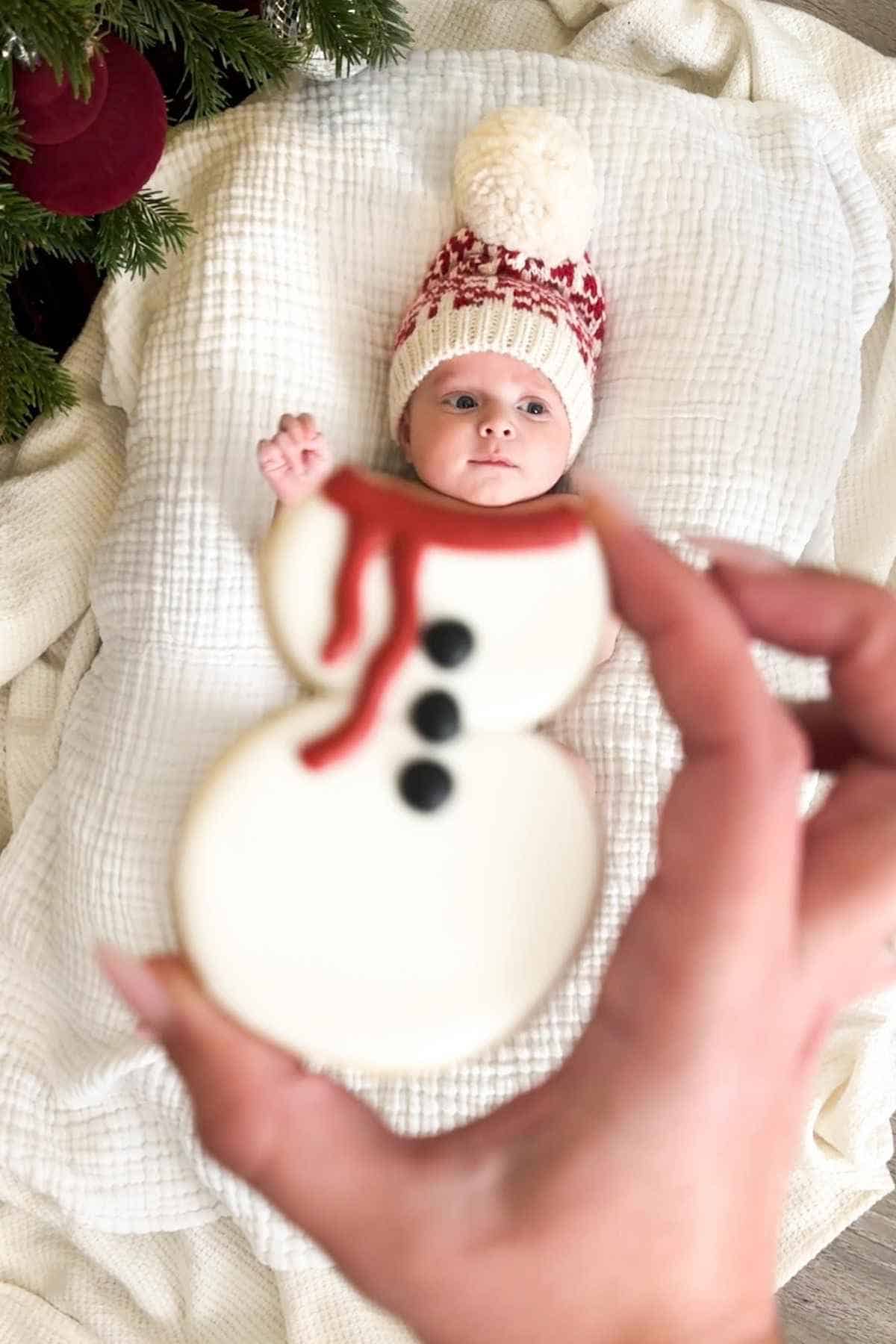 A baby wearing a knitted hat lies on a blanket near a Christmas tree.
