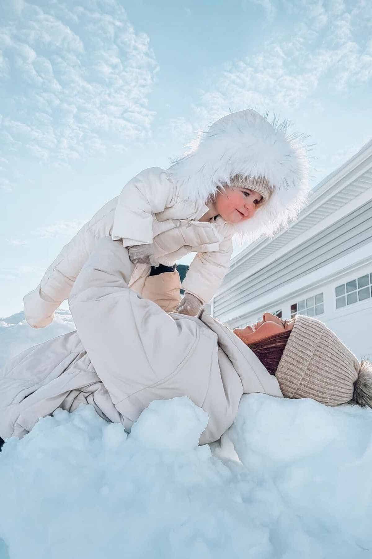 Adult lying in the snow, playfully lifting a child wearing a white snowsuit and fluffy hood under a clear sky.