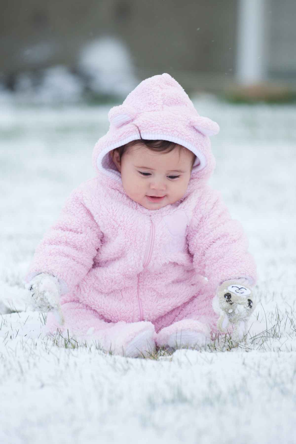 A baby in a pink winter onesie sits on snow-covered grass, looking down.