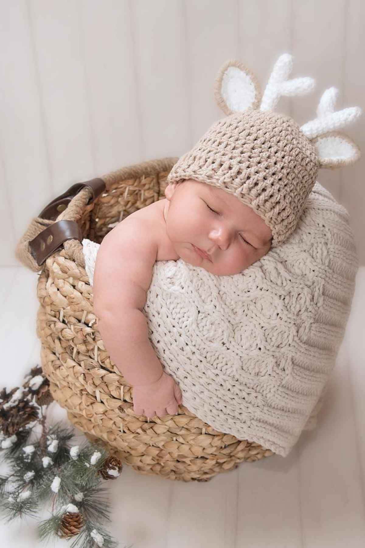 A baby sleeps in a wicker basket, wearing a knitted hat with antler decorations and a white knitted blanket.