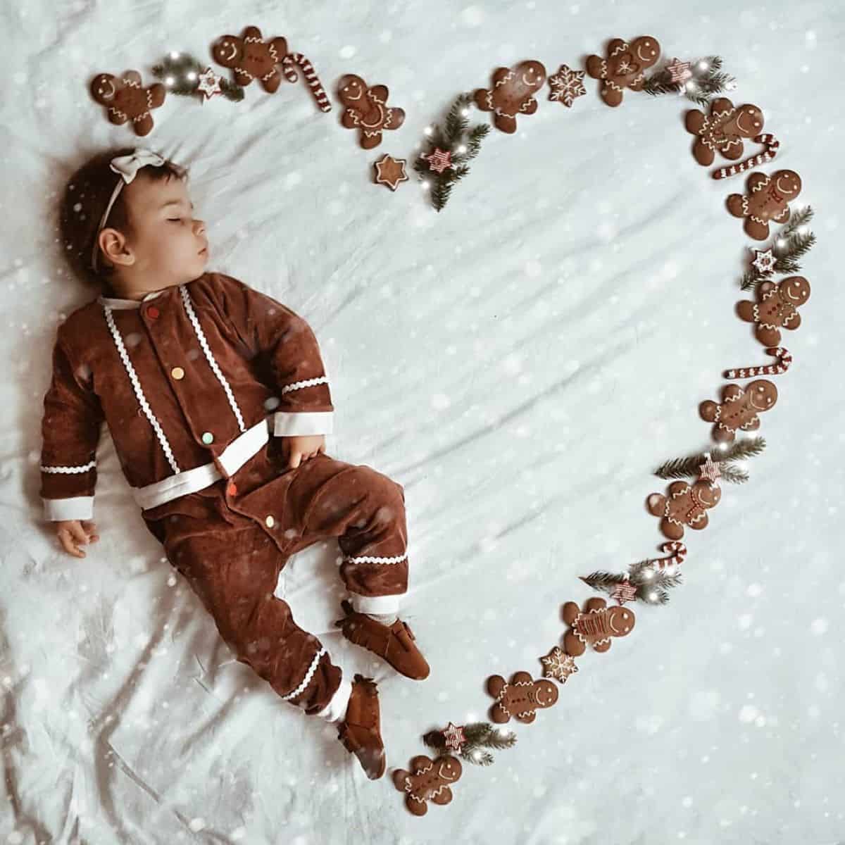 A baby in a gingerbread-themed outfit sleeps on a blanket, surrounded by gingerbread cookies arranged in a heart shape.