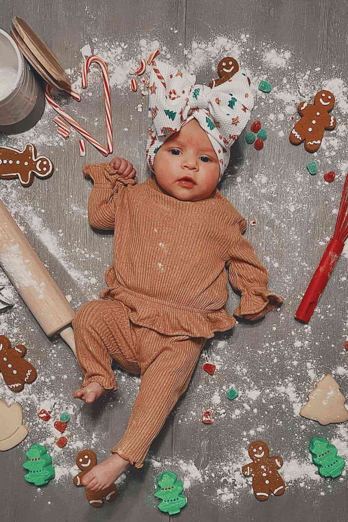 Baby lying on a flour-dusted surface wearing a brown outfit and patterned head wrap, surrounded by gingerbread cookies, candy canes, and baking items.
