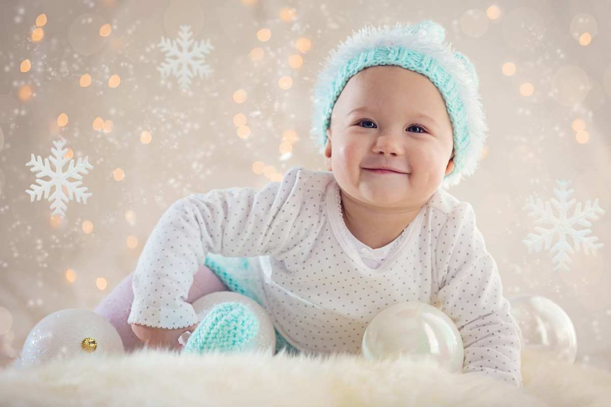 A baby in a white polka dot outfit and blue knit hat smiles while surrounded by snowflakes and festive lights.