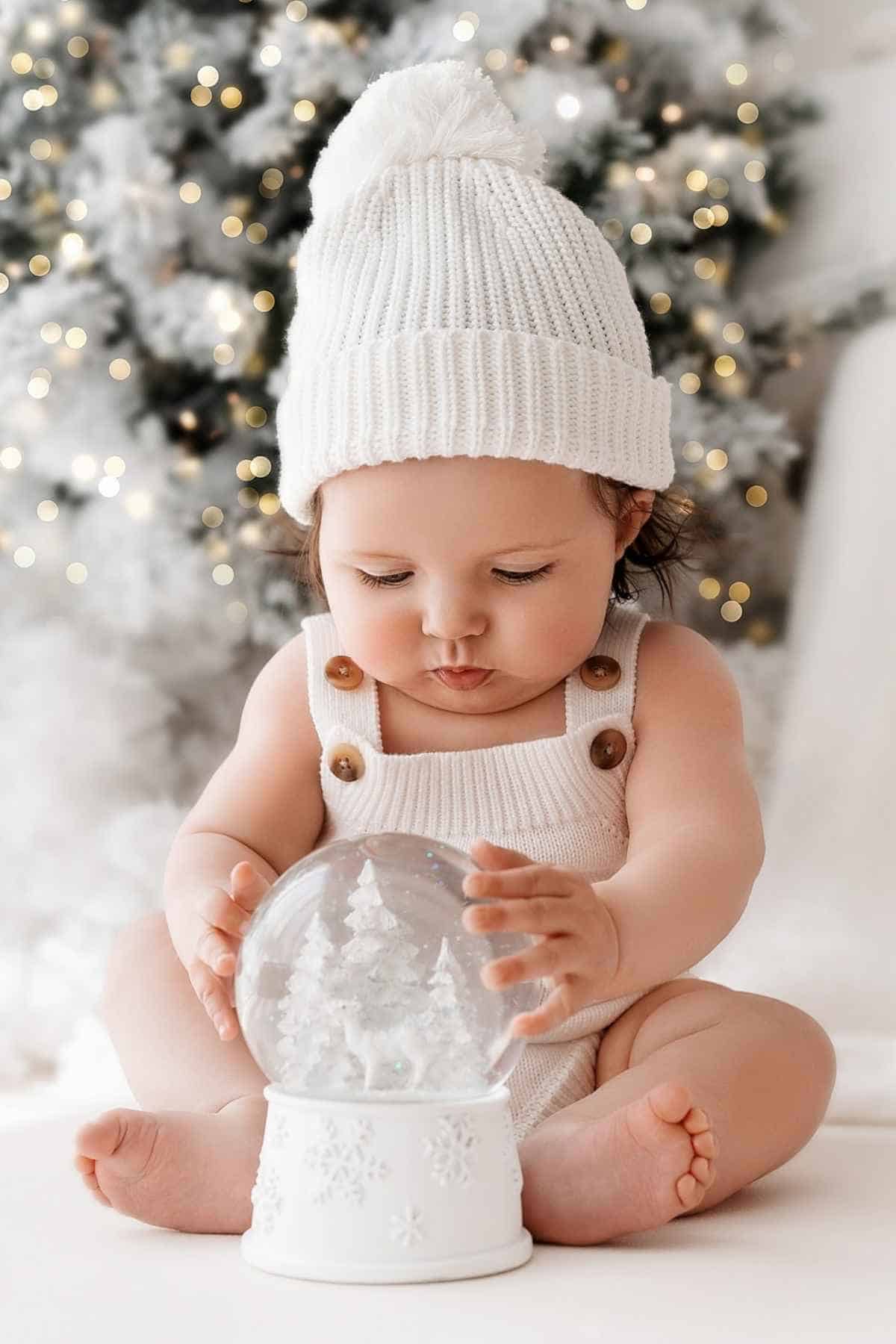 A baby in a white knitted hat and outfit sits on the floor, holding a snow globe with a Christmas tree inside.