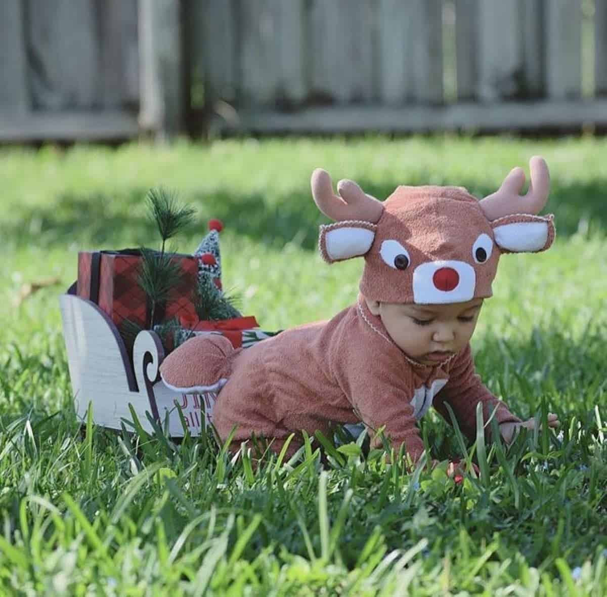 A baby in a reindeer costume crawls on grass next to a small wooden sleigh with wrapped gifts.