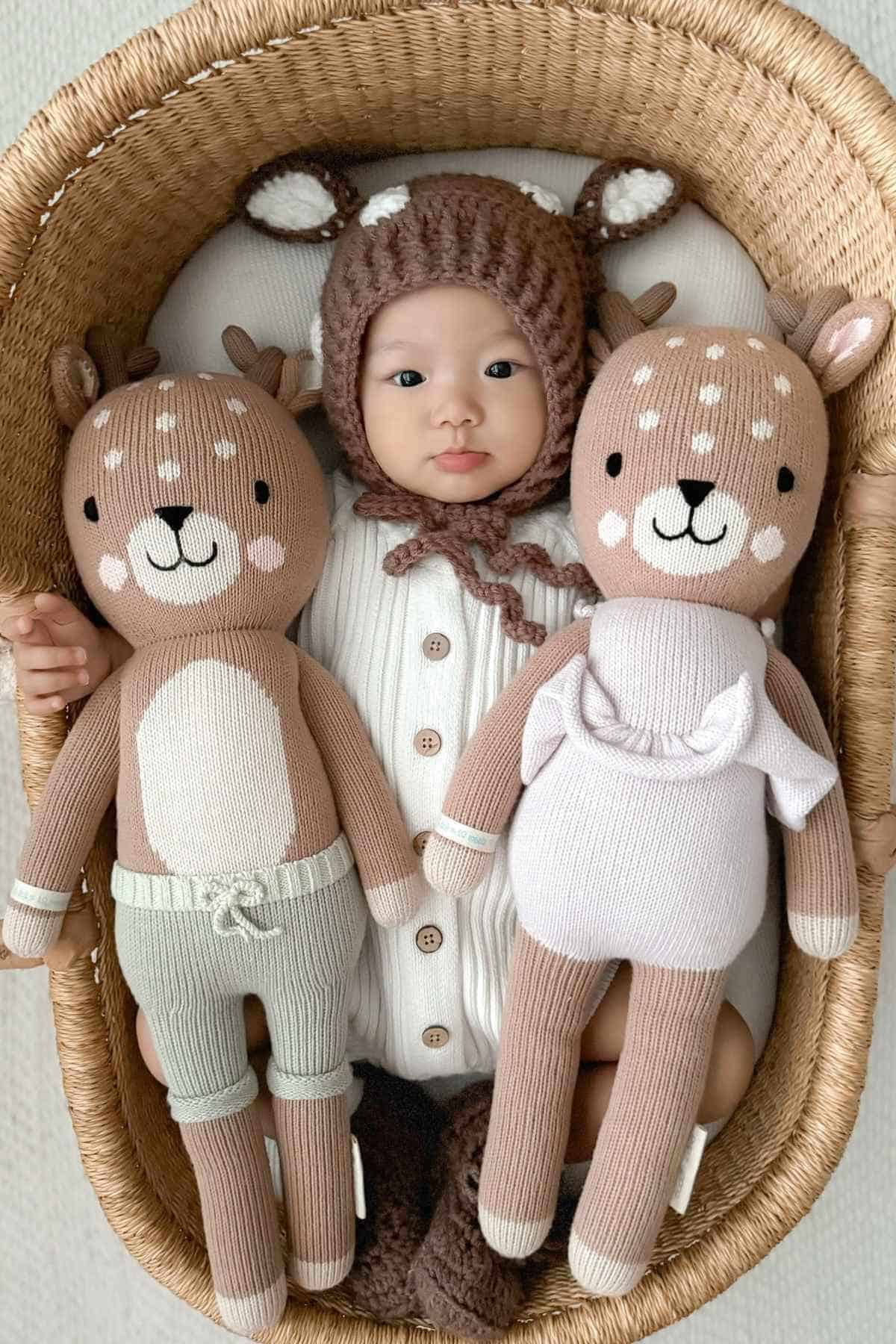 A baby wearing a deer-themed outfit is lying in a basket, holding two stuffed deer toys.