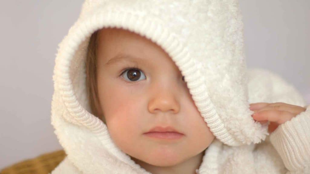 A young child in a fluffy white hooded garment with one eye visible, looking directly at the camera.