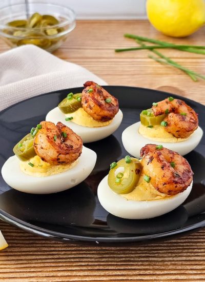 Cajun deviled eggs with shrimp are topped with sliced jalapeños, artfully arranged on a black plate set against a rustic wooden table, accompanied by a refreshing lemon wedge.