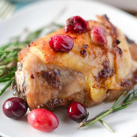A piece of roasted chicken thighs with cranberry sauce, garnished with rosemary, rests elegantly on a white plate.