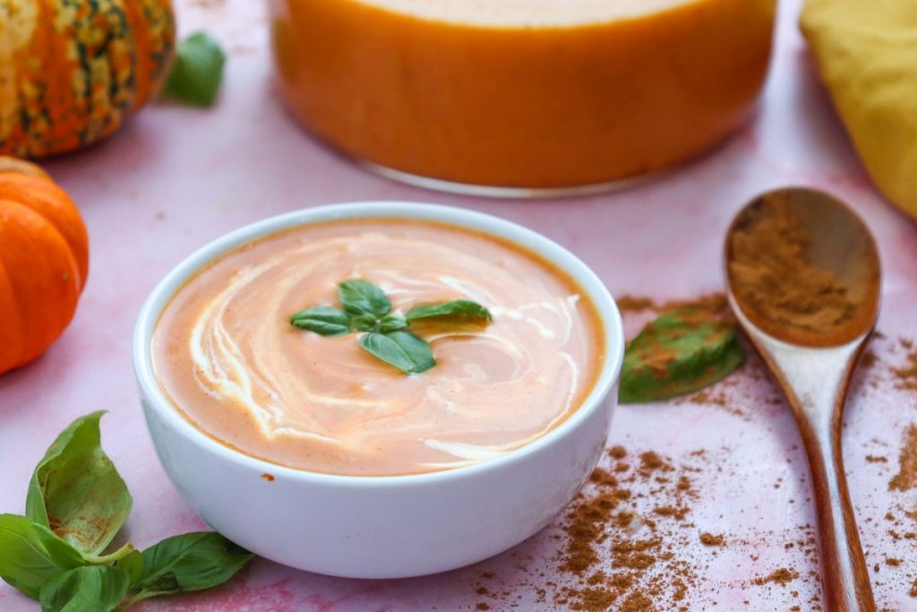 A bowl of pumpkin soup, creamy and garnished with basil leaves, sits invitingly next to a spoon with spices, a whole pumpkin, and a slice of cheese on a light pink surface.