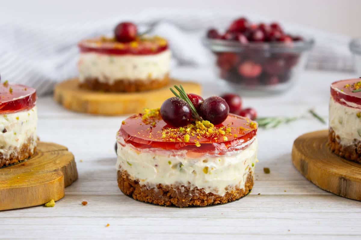 Mini cheesecakes topped with red cranberry jelly and cranberries, garnished with pistachios, on wooden coasters. A bowl of cranberries in the background.