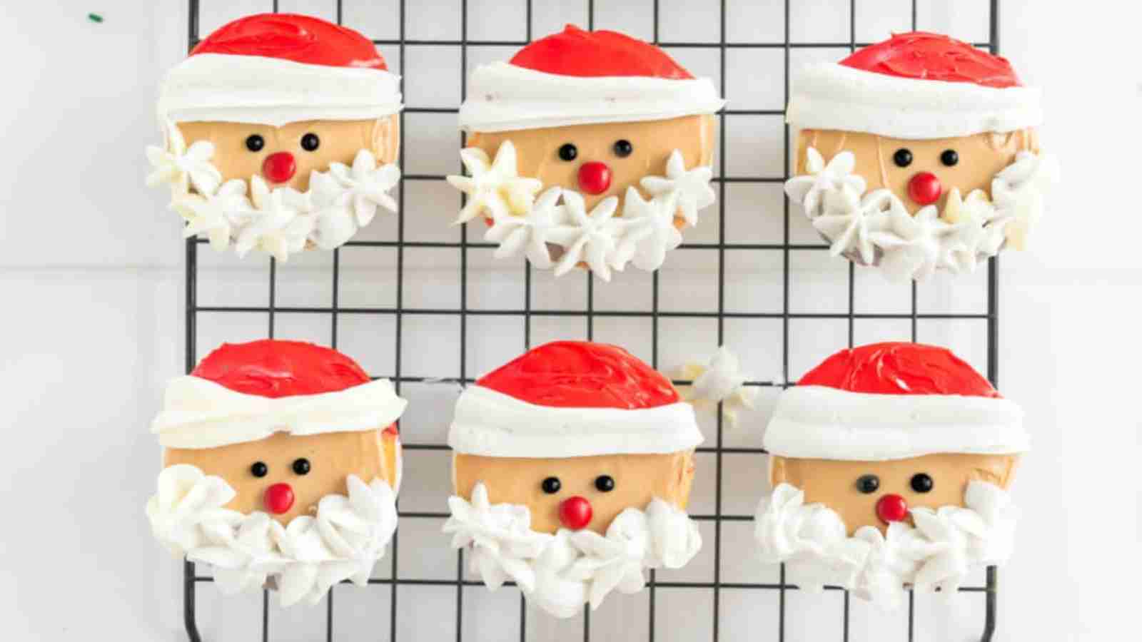 Six Santa-themed cupcakes with red hats, white frosting beards, and black and red candy face decorations on a cooling rack.