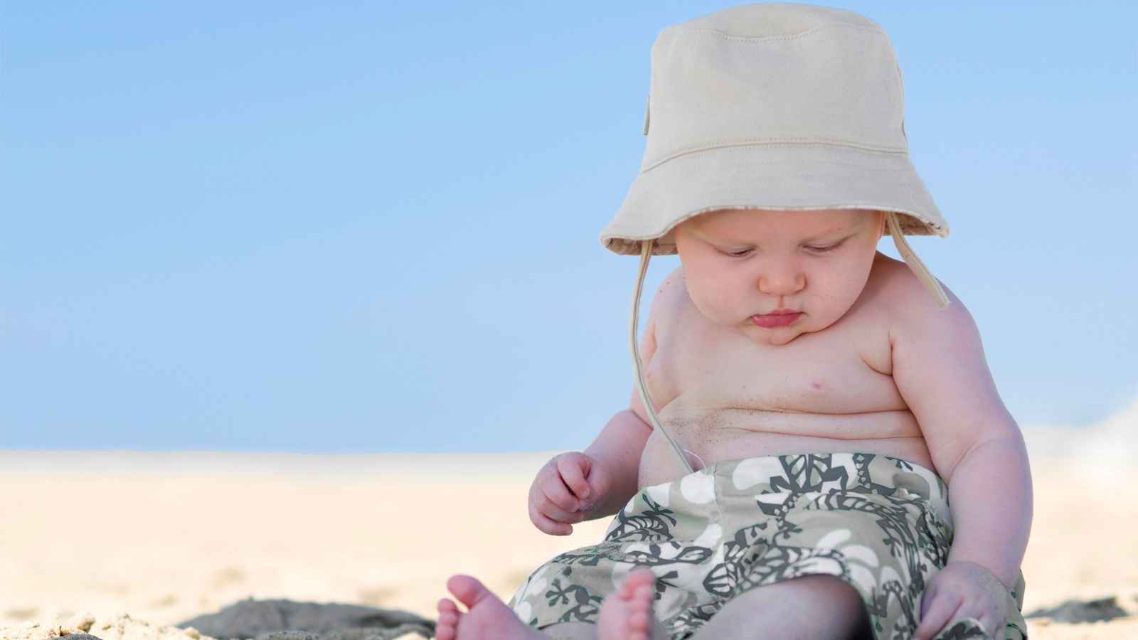 A baby wearing a sun hat and patterned shorts sits on the sandy beach, looking down with a blue sky in the background.