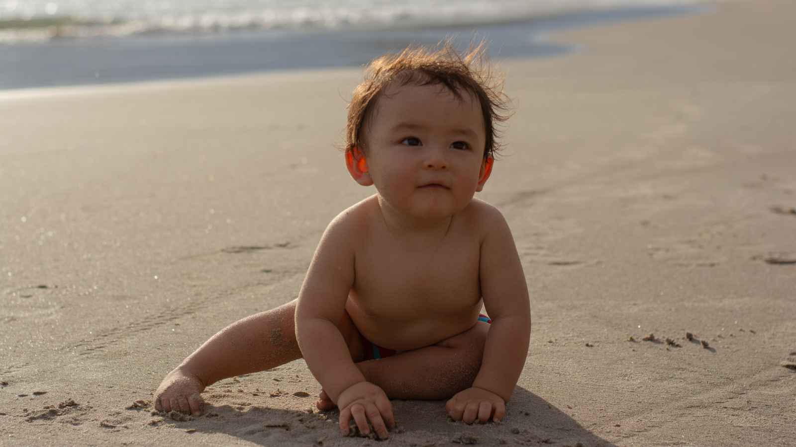 A baby sits on sandy beach near the ocean, gazing forward with a neutral expression.