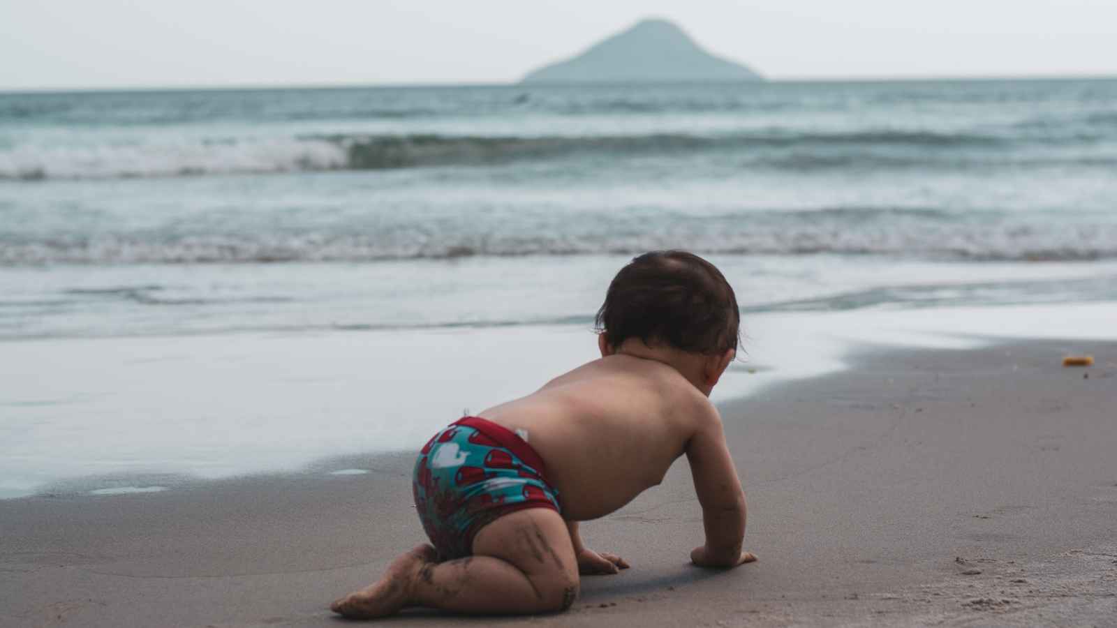 A toddler in swim trunks crawls on a sandy beach towards the ocean, with an island visible in the distance.